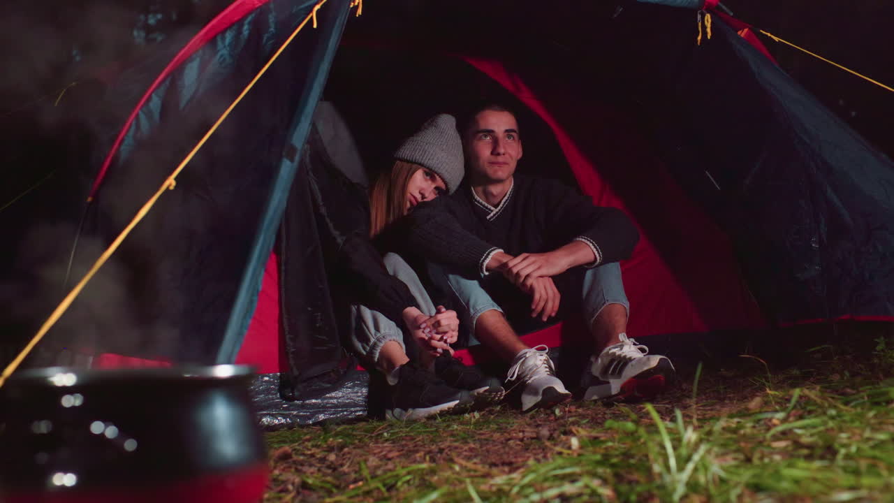 Duo lover seated together at tent entrance during night camping, sharing peaceful moment near steaming pot over campfire, surrounded by quiet forest
