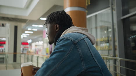 young man sipping warm beverage from paper cup while leaning on counter in modern urban environment, dressed in cozy hoodie and denim jacket, enjoying calm moment in public space