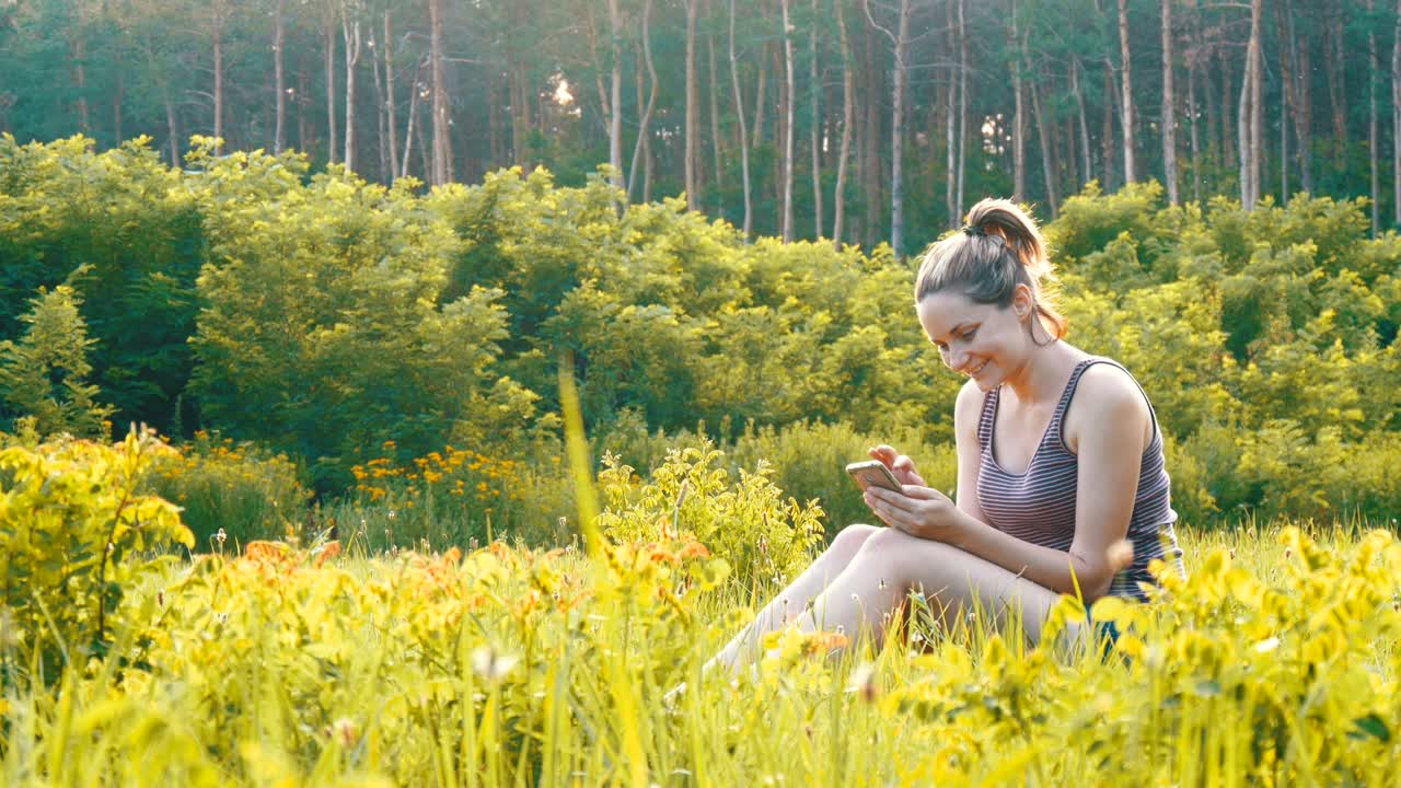 Happy Young Woman Sitting on Green Lawn and Uses Smartphone on Scenic Field at Sunset Background