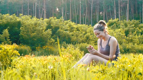 happy young woman sitting on green lawn and uses smartphone on scenic field at sunset background