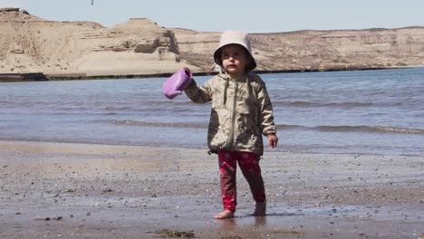 Cute-Little-Girl-Wearing-Bucket-Hat-Walking-And-Holding-A-Plastic-Watering-Can-Toy-At-The-Beach-On-A-Sunny-Summer-Day