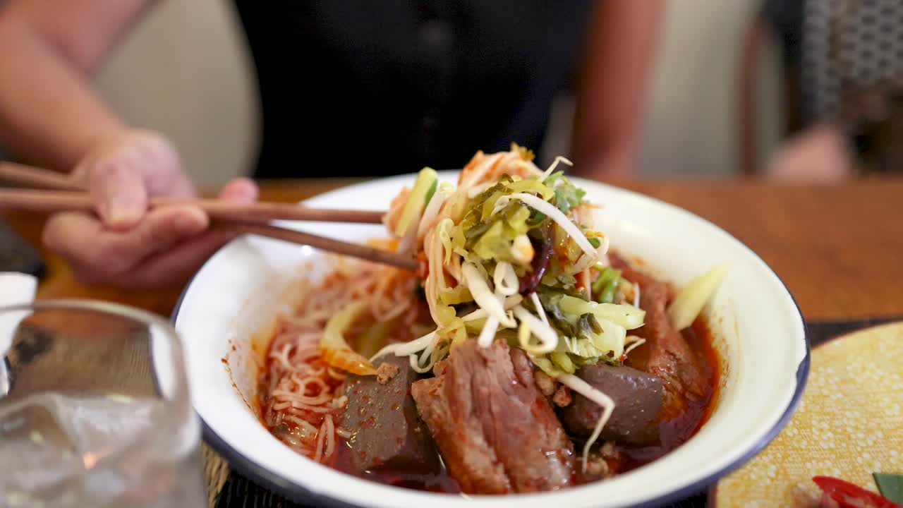 Hand uses chopsticks to lift rice vermicelli noodles from a bowl of Northern Thai curry, highlighting vibrant ingredients in a warmly lit restaurant setting