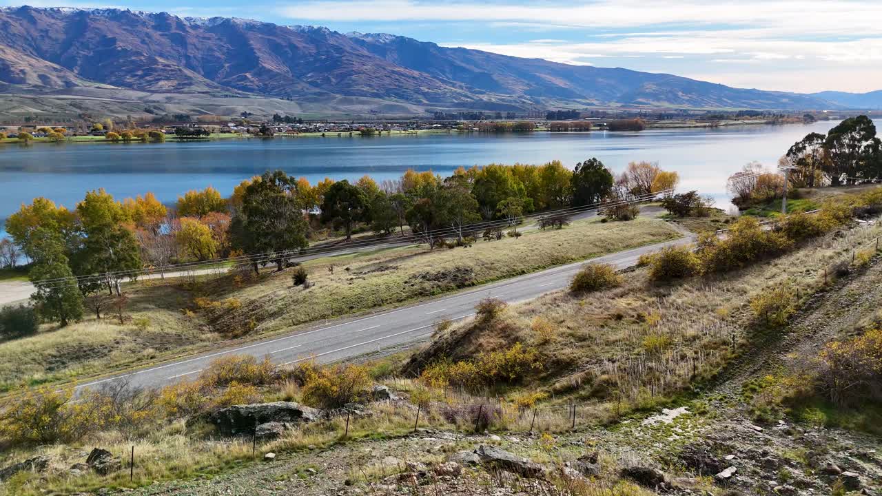 Aerial view of Lake Dunstan with vibrant autumn foliage, showcasing serene landscapes and a passing vehicle on a winding road