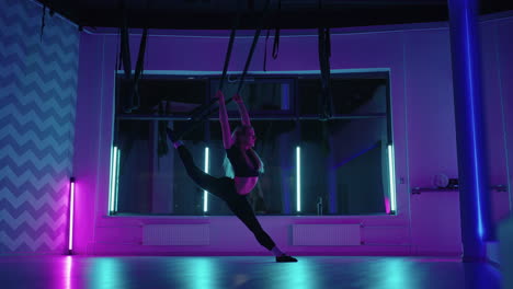 una mujer hace yoga deportivo en el aire en una hamaca a la luz del neón realizando ejercicios y movimientos graciosos de estiramiento y baile en cámara lenta.