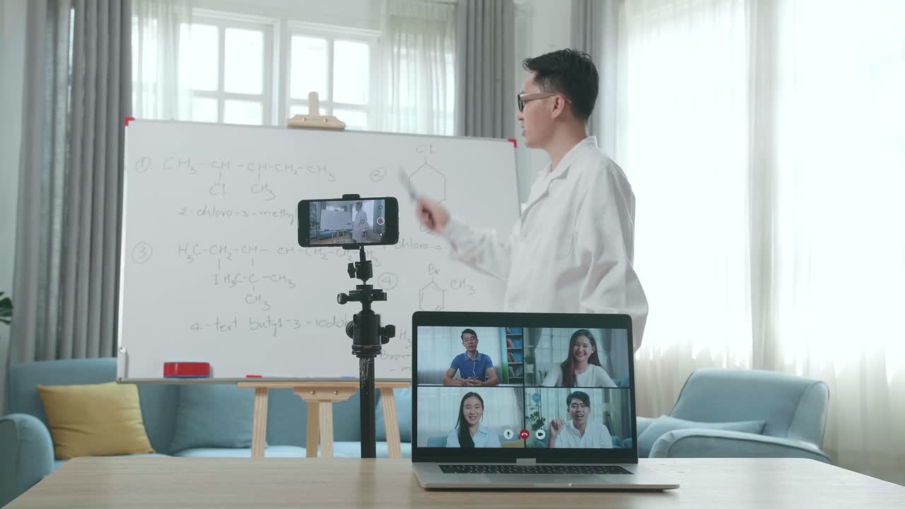 Asian Male Teacher With Glasses Shoots Video By Smartphone Camera And Having Video Call On Laptop While Teaching Chemist At Home