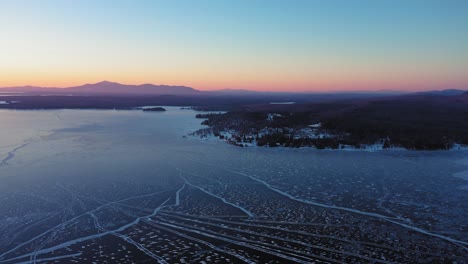 Aerial-view-flying-down-towards-shore-high-above-long-cracks-in-the-ice-of-a-frozen-lake-with-a-mountain-ridge-in-the-distance-silhouetted-by-sunrise