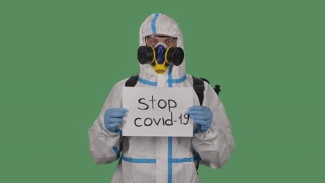 portrait of a man in a protective suit, safety glasses, gloves and a respirator looks into the camera while holding a stop covid 19 sign. green screen, chroma key. slow motion. close up