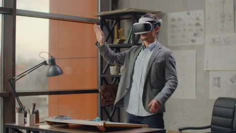 Moderner-Mann,-Architekt-Im-Büro-Mit-Großen-Fenstern,-Steht-In-Einem-Virtual-Reality-Helm-Und-Nutzt-Gesten,-Um-Das-Projekt-Zu-Verwalten,-Ohne-Das-Büro-Zu-Verlassen.-Baukontrolle.-Designprojekt