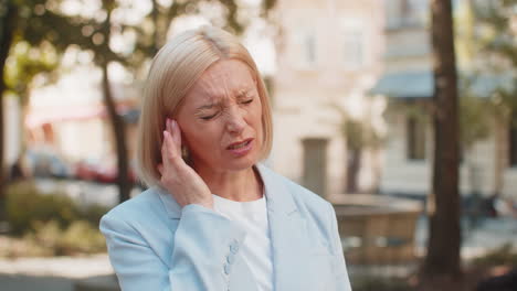 sick mature businesswoman in formal suit suffers from ear pain while standing on city town street