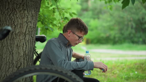 two young siblings rest under a tree, with one drinking water while the other looks into the distance, they are taking a break from a bike ride, with bicycles parked nearby