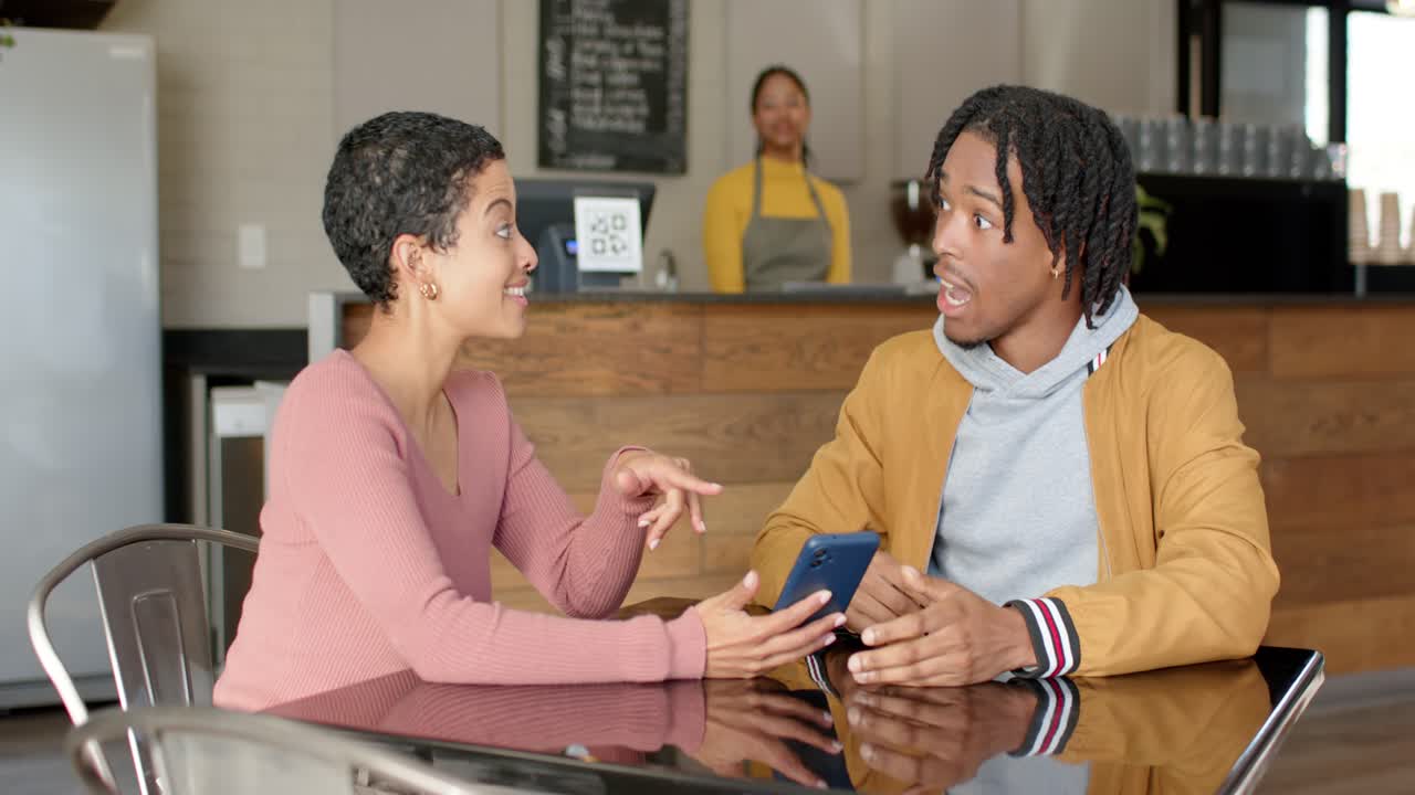 Woman retrieving smartphone, showing content to partner at cafe table, sparking surprised reaction