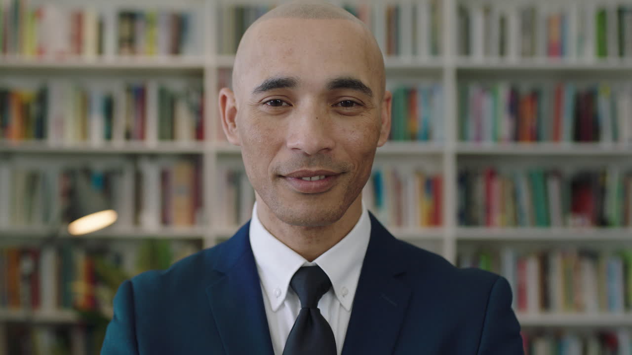 ritratto ravvicinato di un uomo d'affari ispanico calvo che guarda la fotocamera in piedi scaffale della biblioteca sullo sfondo
