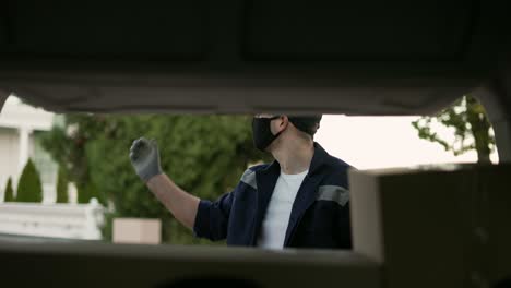 Delivery-service.-Delivery-man-in-a-protective-mask-and-gloves-near-a-car-putting-cardboard-box-into-the-trunk.-Man-closing