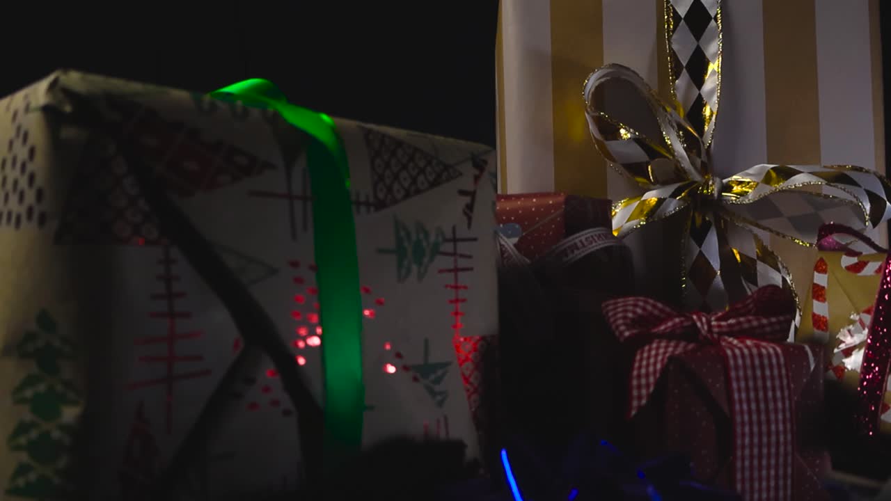 Closeup view sweeps along row of wrapped Christmas presents adorned with ribbons and bows against black background under studio lights. Glossy rosettes fall in slow motion, bouncing off the gift boxes