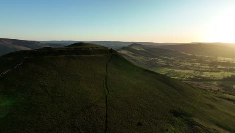 Wide-aerial-orbit-of-Mam-Tor-and-the-Great-Ridge-at-sunrise-in-the-Peak-District,-UK-with-silhouettes-of-people-enjoying-the-view-from-the-trig-point