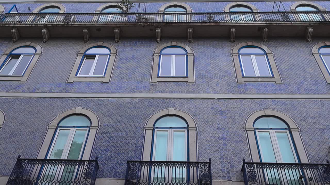 Lenta ripresa dal basso di un edificio residenziale storico di Lisbona caratterizzato da motivi di piastrelle azulejo blu, finestre ad arco ed eleganti balconi in ferro battuto in una morbida luce diurna