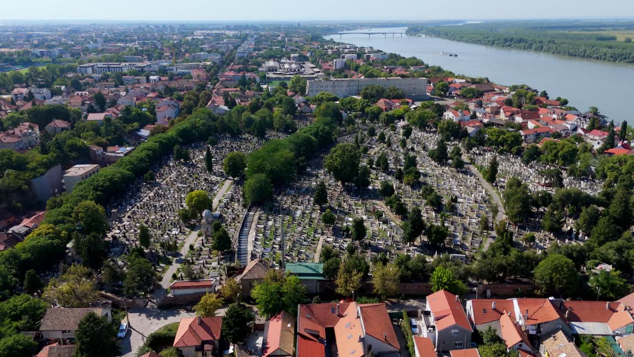 Aerial drone view of Zemun Cemetery in Belgrade, Serbia, overlooking the Danube River. Rows of tombstones surrounded by trees and historic architecture under clear daylight