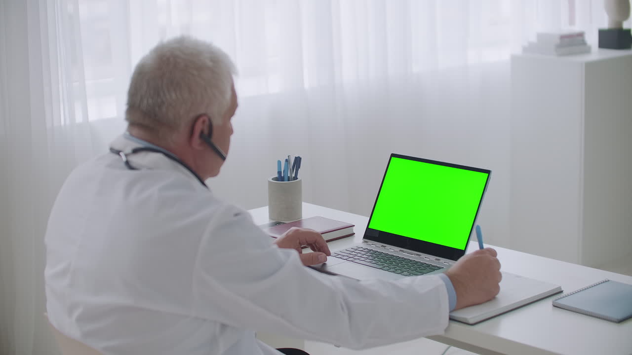 un médico de edad avanzada está escuchando un seminario web o un paciente en una computadora portátil con pantalla verde para la tecnología de llave de croma escribiendo notas en un cuaderno