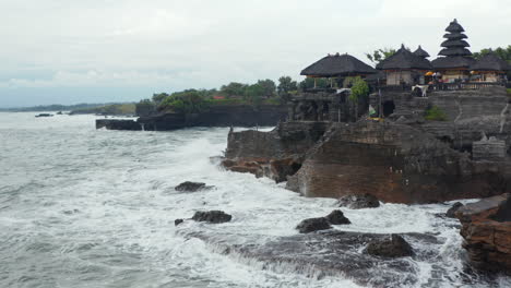 Low-aerial-view-of-strong-ocean-waves-crashing-into-dark-rocky-cliff-with-Tanah-Lot-temple-in-Bali,-Indonesia.-Famous-tourist-destination-in-a-bad-weather