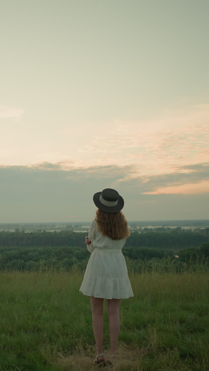 하 드레스와 모자를 입은 한 여인이 해가 지는 동안 조용한 호수 의 풀에서 사려 깊게 서 있습니다. 그녀는 자연의 조용한 설정에서 조용하고 반영적인 순간을 포옹하여 멀리 쳐다보고 있습니다.