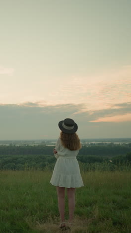 una mujer con un vestido blanco y un sombrero se encuentra pensativa en un campo cubierto de hierba junto a un lago tranquilo al atardecer. ella mira a la distancia, abrazando el momento tranquilo y reflexivo en el entorno sereno de la naturaleza
