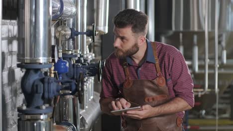 young male brewer wearing a leather apron supervise the process of beer fermentation at a modern brewery factory