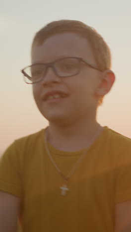 a young boy wearing glasses and a yellow shirt walks in a sunlit grassy field, smiling shyly while moving his hands playfully. the warm glow of the sunset enhances the serene and joyful atmosphere