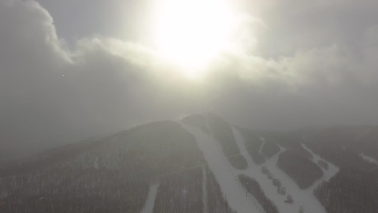 Bright sun shining through fluffy clouds over snow covered ski slopes and a serene forest in Orford, Quebec, Canada, creating a misty and atmospheric winter landscape