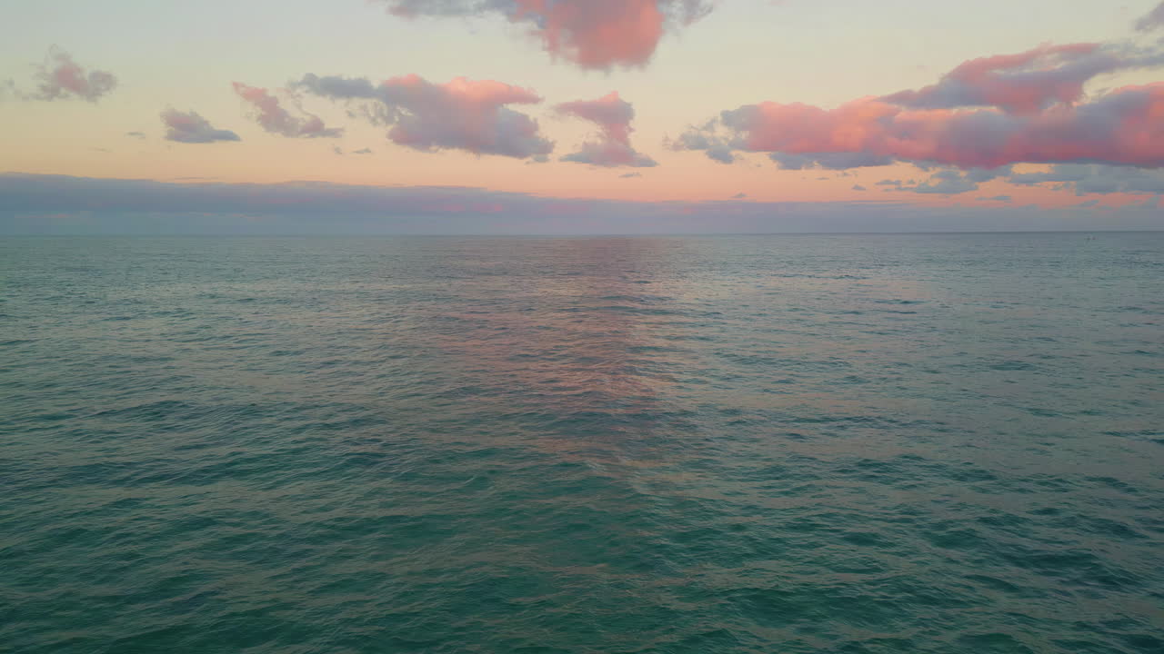 Drone soft ocean sunset bringing pink hue to clouds over calm tranquil ocean.