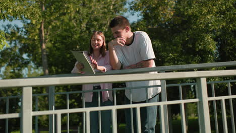 girl shows boy something on her tablet with light reflecting on her face, both smiling as he points at the screen with hand tattoo, set against a lush backdrop of greenery and trees