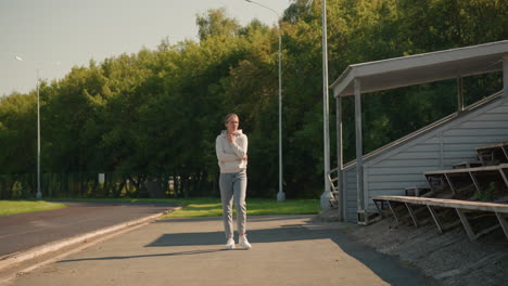 young woman in sporty attire with white sneakers walks near a stadium on a sunny day, passing by electric poles and surrounded by lush green trees