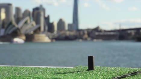 Primer-Plano-De-Un-Club-De-Golf-Golpeando-Una-Pelota-En-Cámara-Súper-Lenta-Con-La-Ciudad-De-Sydney-En-El-Fondo