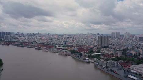Aerial-shot-along-Saigon-river-port-with-navy-ships,-tourist-boats-and-urban-sprawl