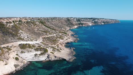 Drone-Aéreo-Filmado-En-4k-De-La-Hermosa-Costa-Rocosa-De-La-Isla-Balear-De-Mallorca-Con-Aguas-Cristalinas-Y-Turquesas-Junto-A-Los-Acantilados-Con-Barcos-Y-Yates-De-Vela-En-El-Mar-Mediterráneo