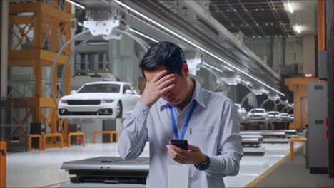 trabajador profesional masculino asiático de pie con su teléfono inteligente en la fábrica de automóviles ev, instalación industrial de alta tecnología, está asintiendo con la cabeza con decepción