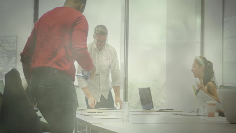 coworkers leaning over table, adjusting documents, viewing tablet overlays in business meeting