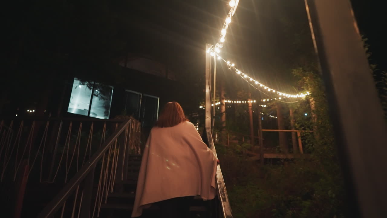 una mujer envuelta en una manta sube las escaleras. una mujer regresa a la residencia después de un paseo nocturno recibida por el cálido resplandor de bombillas decorativas que iluminan la escalera