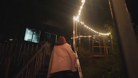 lady wrapped in blanket ascends stairs. woman comes back to residence after evening stroll greeted by warm glow of decorative bulbs illuminating staircase