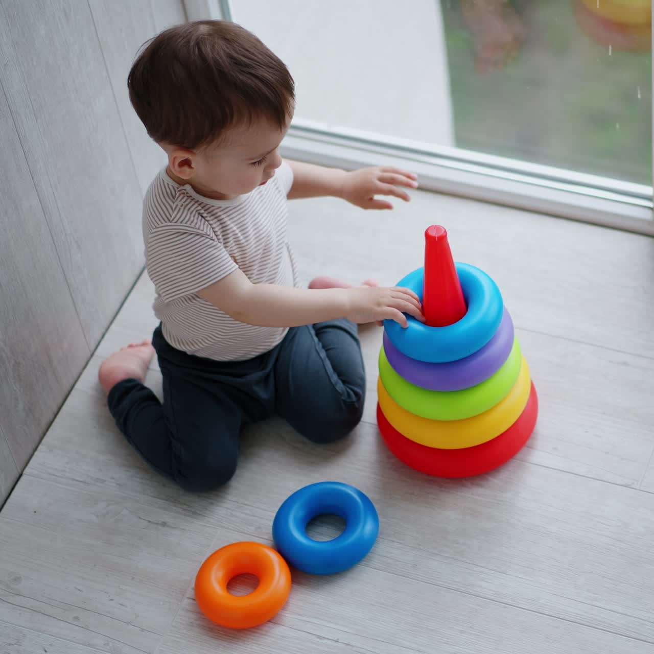 Adorable Caucasian toddler sits on his knees on the floor. Child constructs the pyramid on the balcony. View from top