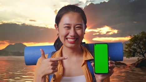 Close Up Of Asian Female Hiker With Mountaineering Backpack Smiling And Pointing To Mock Up Green Screen Smartphone While Standing At A Lake During Sunset Time