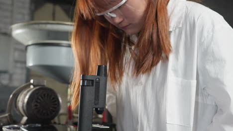 lab scientist examines sample through microscope, her hair cascading over her shoulder, while tapping on a tablet in a mechanical shop filled with tools and equipment