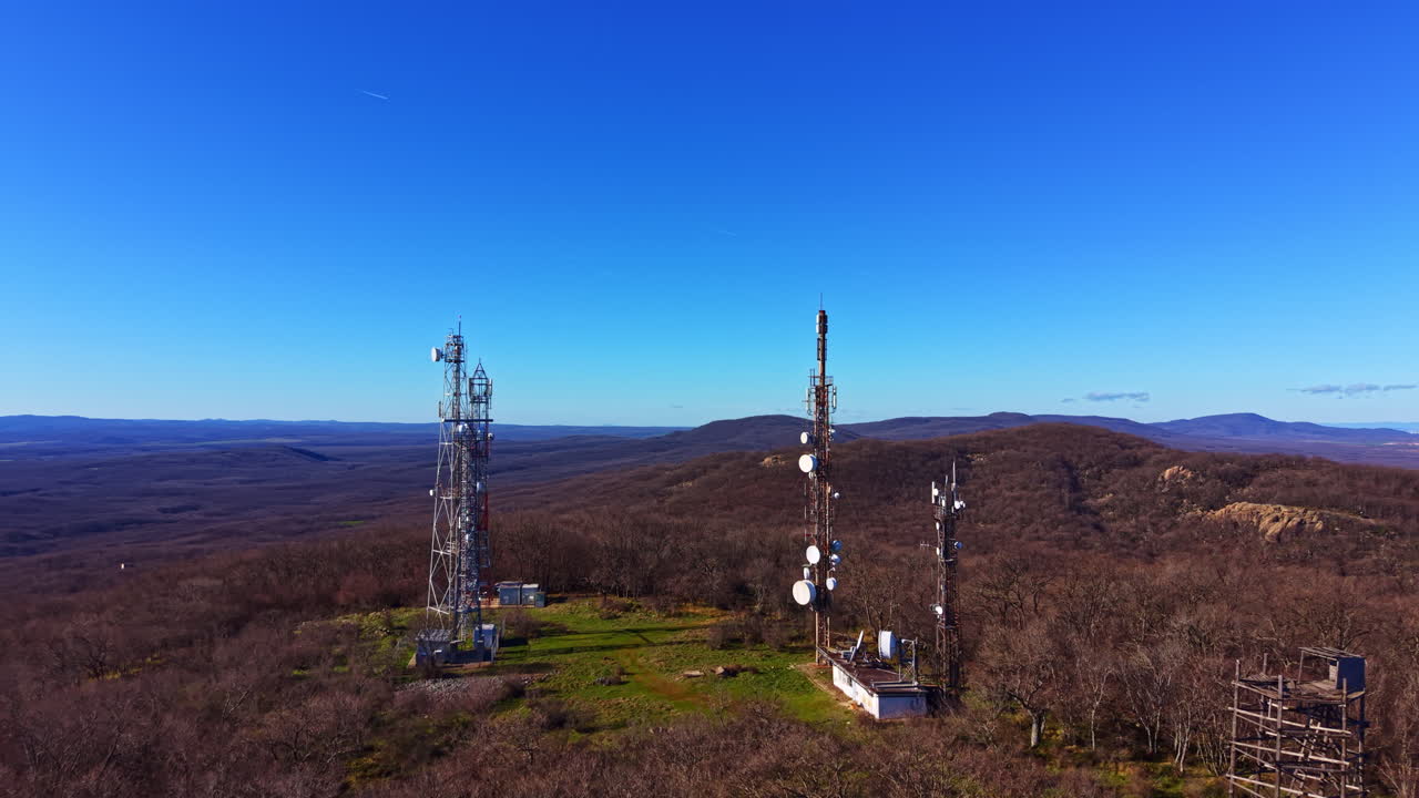 Communication towers stand on a hill in a rural area under a clear blue sky during daytime in a mountainous region