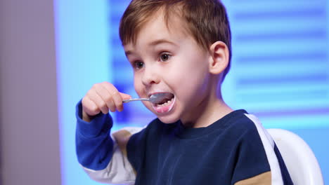 cute dark-haired toddler wearing a sweater eats dairy from spoon. lovely baby enjoying yogurt. close up.