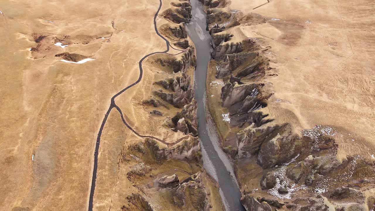 Top-down view of Fjaðrárgljúfur Canyon in Iceland, revealing winding paths, sheer cliffs, and a serpentine river slicing through the rugged golden landscape.