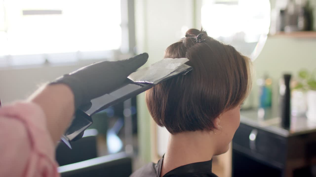 Hands of caucasian male hairdresser highlighting female client's hair at salon, in slow motion