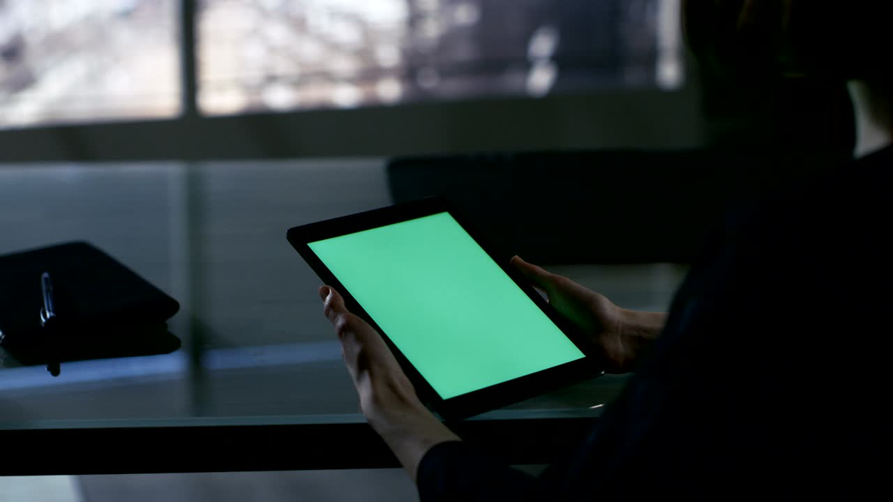 sobre la vista del hombro de la mujer de negocios sentada en su escritorio y usando una tableta con pantalla verde. la gran ciudad se ve por la ventana.