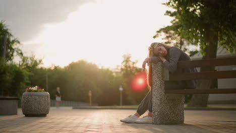 lady seated at the edge of a bench during sunset, resting her head on the side, creating a serene and reflective moment, as sunlight casts a soft glow around her with greenery in the background