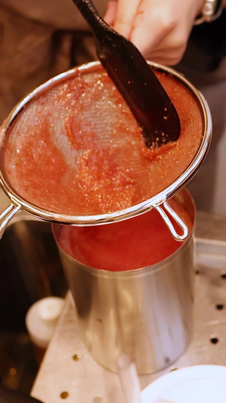 Primo piano di mani che usano una spatola per filtrare purea di frutta rossa in un contenitore di acciaio inossidabile.