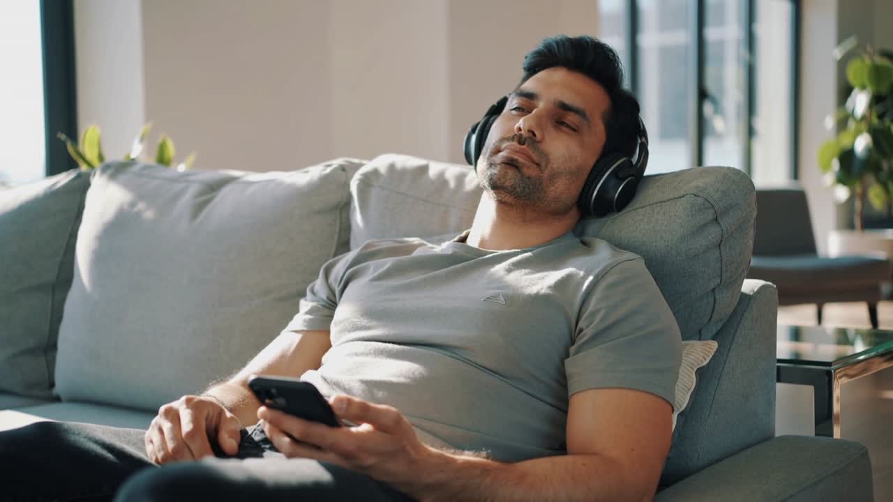 Uomo in maglietta grigia che tocca play sul telefono, reclinato sul divano, rilassandosi mentre ascolta con le cuffie.