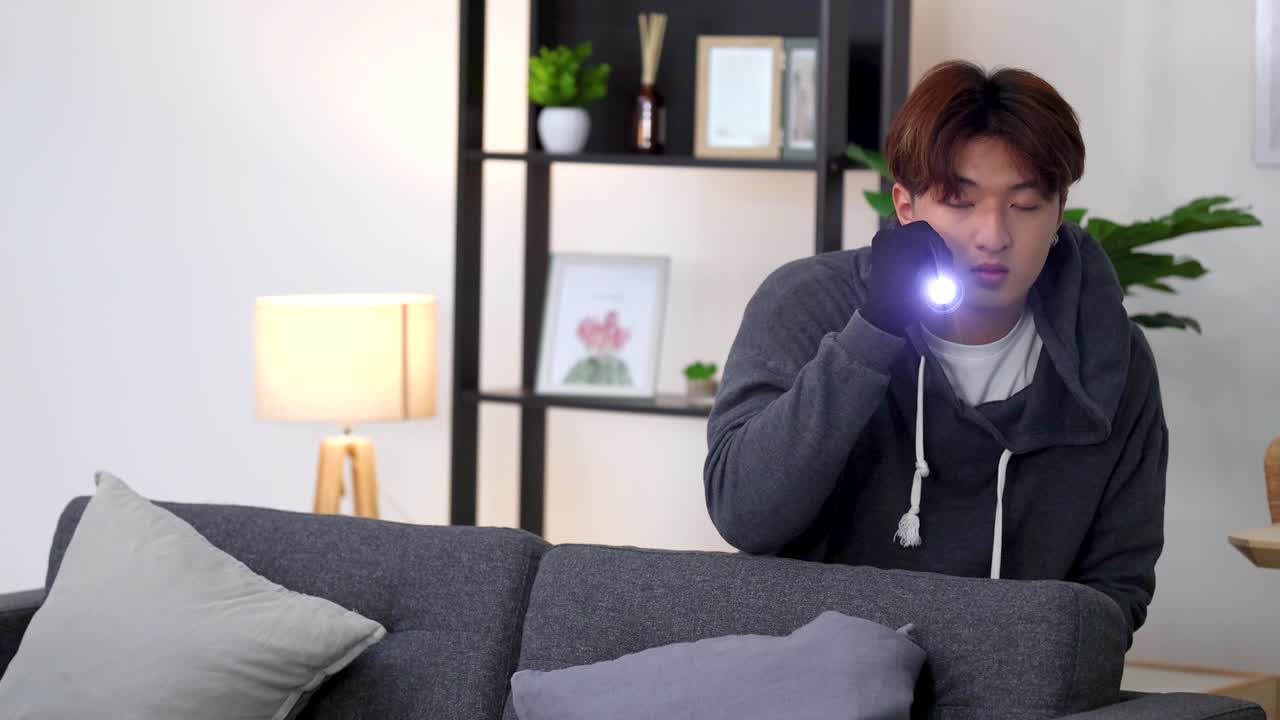 A suspicious man wearing gloves uses a flashlight while searching a living room at night